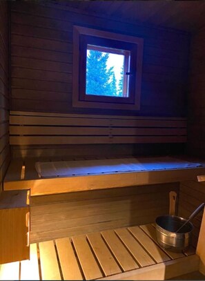 Ein Saunaraum mit einer Sitzbank, einem kleinen Fenster und einem Metallbehälter.