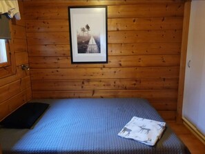 Une petite chambre avec un lit simple, des murs en bois, un tableau encadré au mur et une fenêtre avec des rideaux.