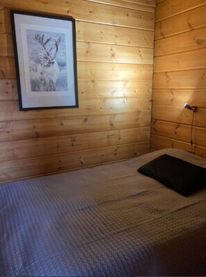 Ein Zimmer mit Holzwänden, einem gerahmten Bild eines Rehs, einem Bett mit strukturierter Decke und einem Nachttisch mit einer Lampe.