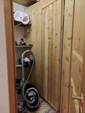 Ein kleiner Raum mit einer Holztür, einem Staubsauger und einem Regal mit verschiedenen Gegenständen.