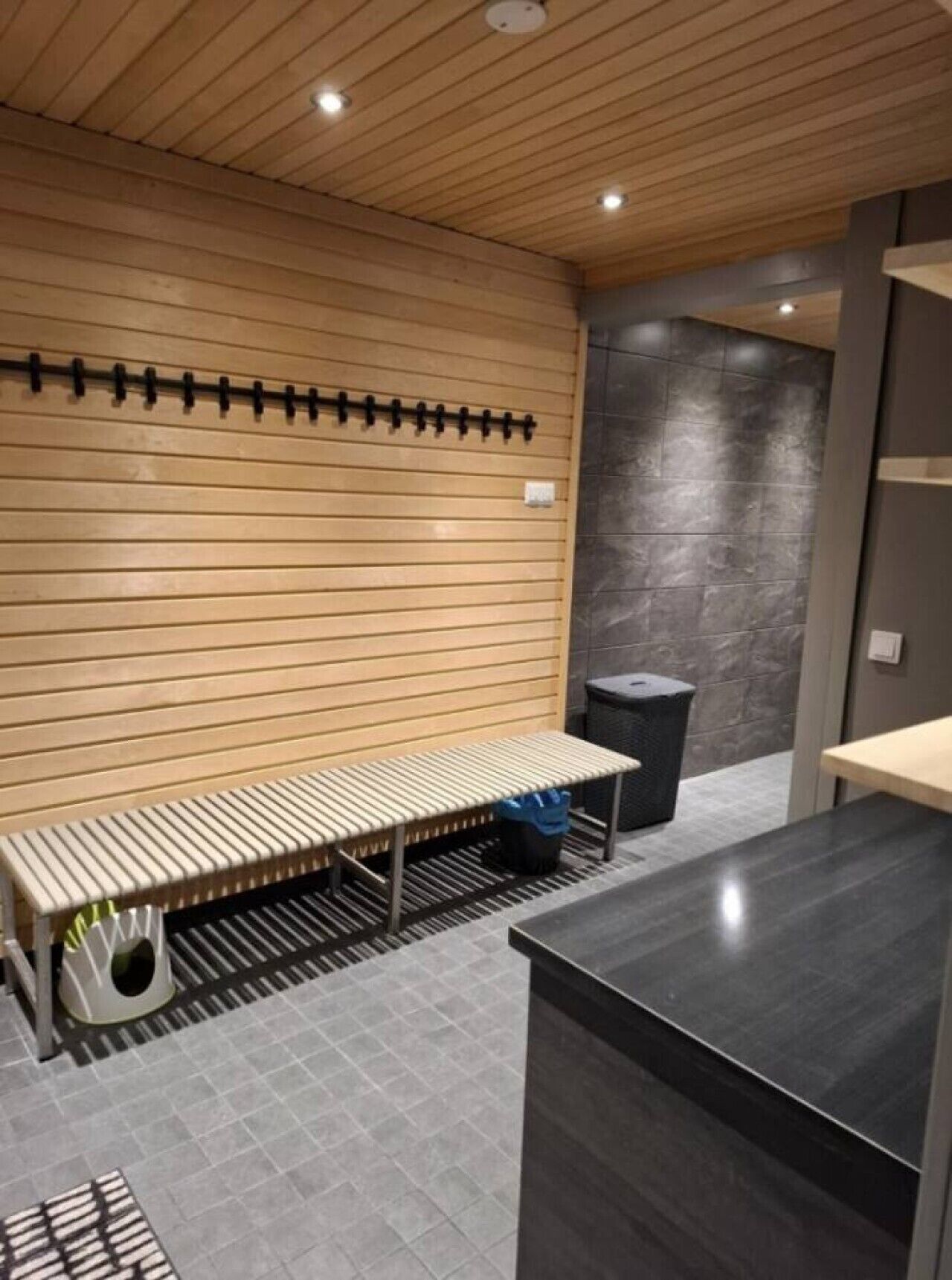Une salle de bain moderne avec un banc en bois à lattes, une poubelle noire et un sol carrelé de couleur grise.