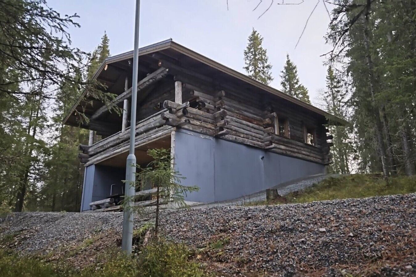 A log cabin with a steeply pitched roof, surrounded by trees and a gravel path.