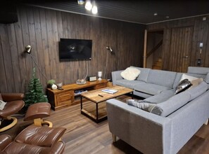 A modern living room with a grey sofa, leather chairs, a wooden coffee table, a small Christmas tree, and a flat-screen TV mounted on the wall. - Rukapirtti | Spacious 5BR Chalet with Sauna (Ruka)