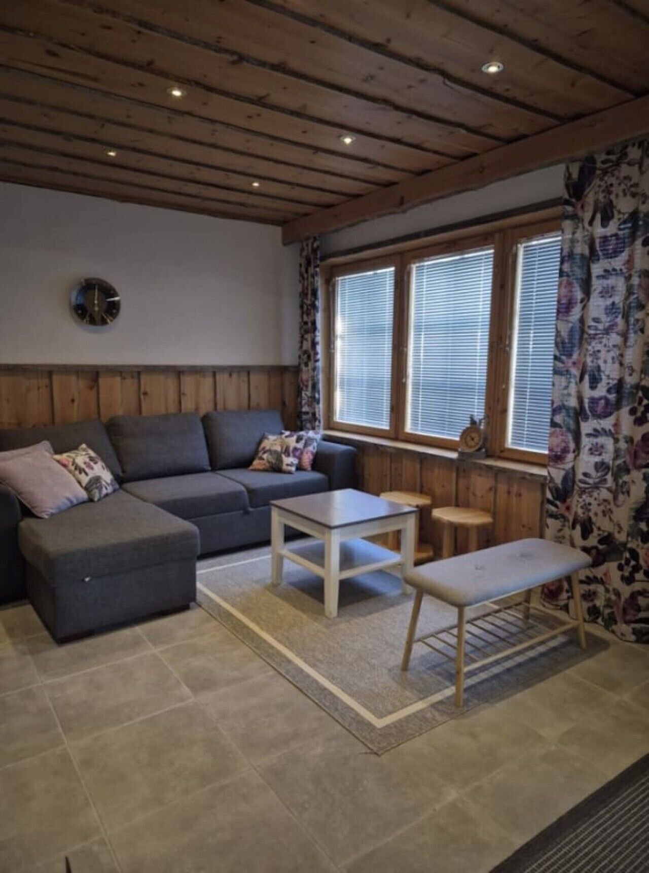 A cozy living room with a sofa, ottoman, coffee table, and bench. There is a window with blinds and a clock on the wall.