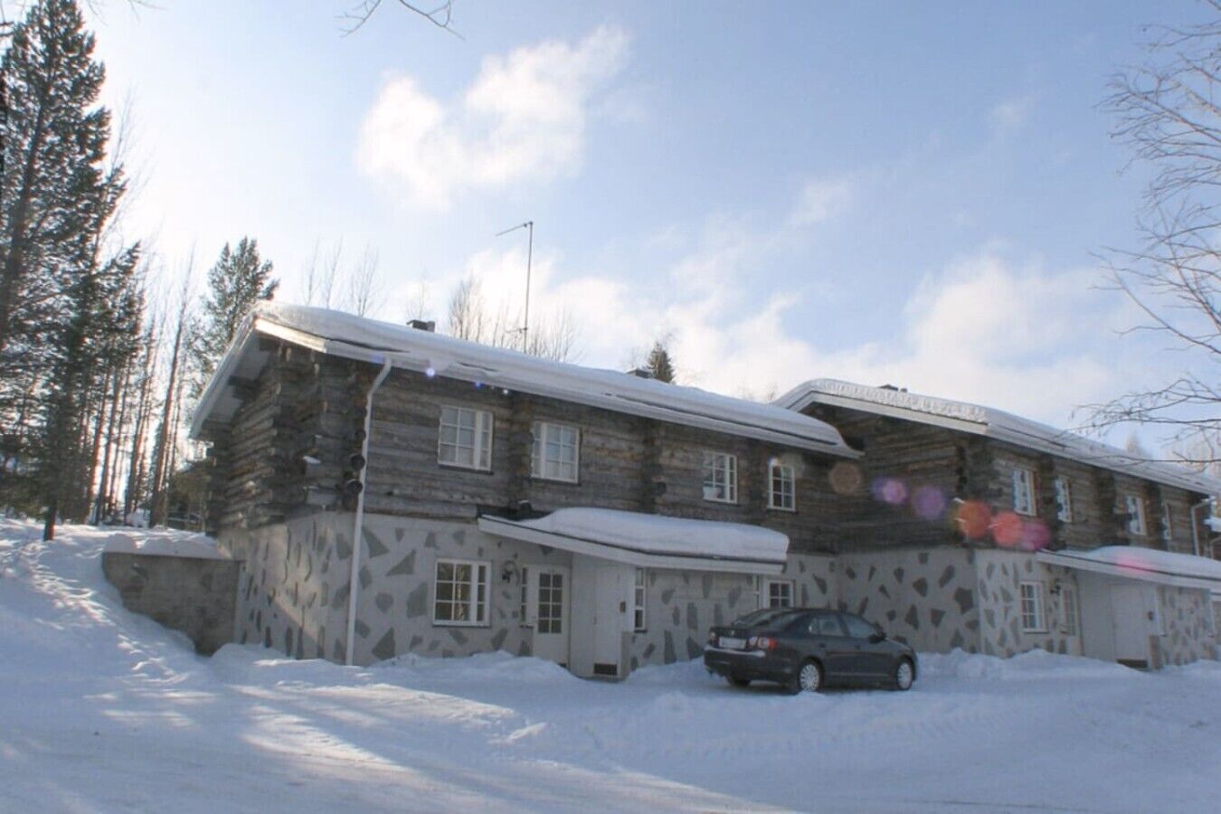 A multi-story log cabin with a snow-covered roof, surrounded by a snowy landscape and trees.