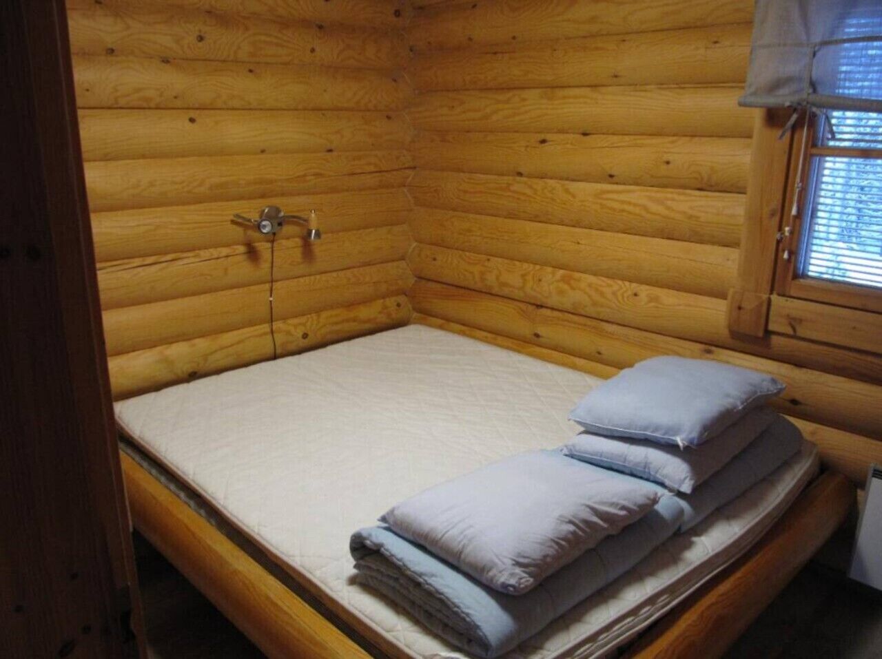 A wooden cabin room with a bed, a window with blinds, and a small heater.