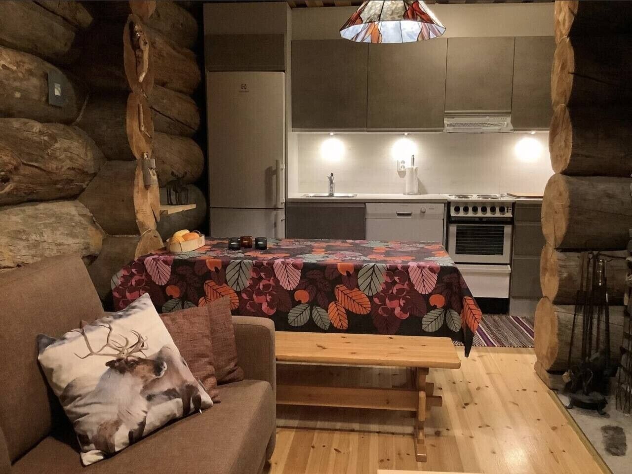 L’intérieur chaleureux d’un chalet avec un banc en bois, un canapé agrémenté d’un coussin à motif de cerf, une table recouverte d’une nappe à fleurs et un coin cuisine équipé d’une cuisinière et d’un réfrigérateur.
