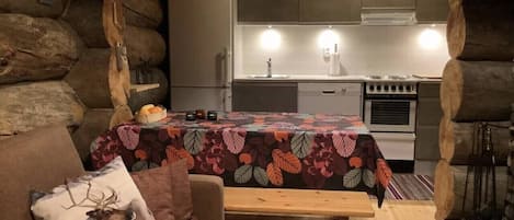 A cozy cabin interior with a wooden bench, a sofa with a deer pillow, a table with a floral tablecloth, and a kitchen area with a stove and refrigerator.