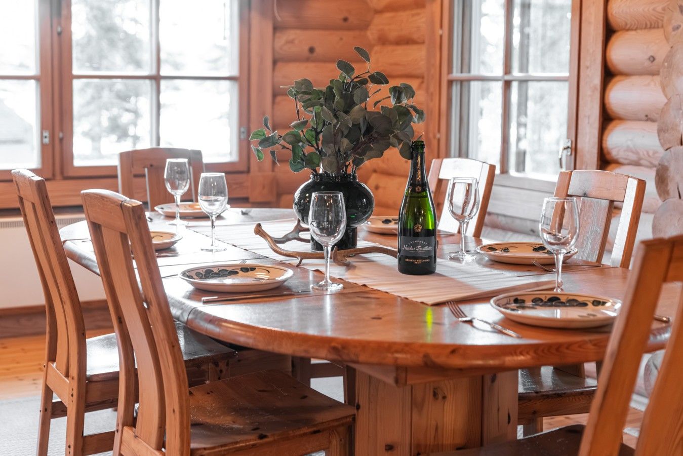 Una mesa de comedor de madera dispuesta con platos, vasos y una botella de vino, rodeada de sillas de madera.