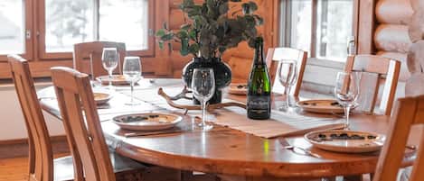 A wooden dining table set with plates, glasses, and a bottle of wine, surrounded by wooden chairs.