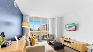 Un salon moderne comprenant un canapé, une table basse et un téléviseur, et offrant une vue sur la ville grâce à de grandes fenêtres.