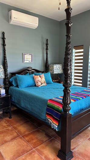 A four-poster bed with a blue bedspread and pillows, a nightstand with a lamp, a wall-mounted air conditioner, and a window with blinds.