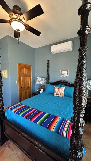 A four-poster bed with a blue bedspread and colorful pillows, a ceiling fan, and an air conditioning unit on the wall.