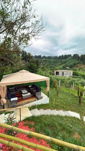 A covered outdoor seating area with a table and chairs, surrounded by greenery and a view of a hilly landscape.