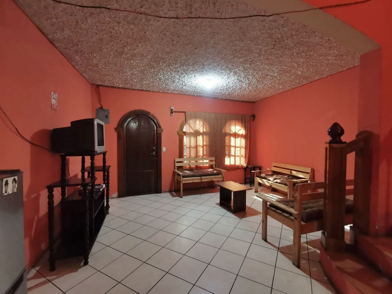 Quarto com piso de azulejos, porta de madeira, televisão sobre um suporte e dois bancos de madeira.