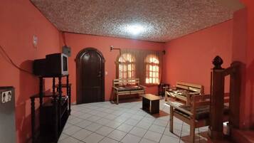 Quarto com piso de azulejos, porta de madeira, televisão sobre um suporte e dois bancos de madeira.