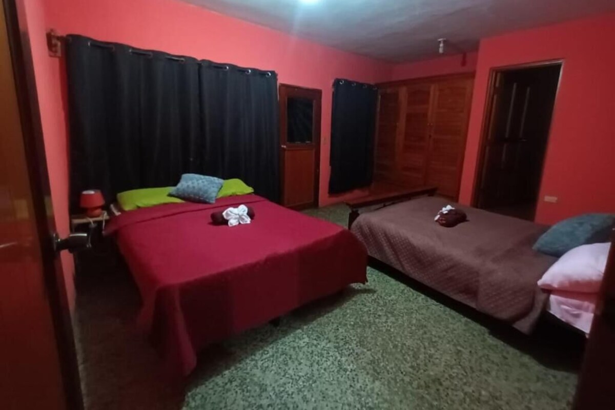Ein Zimmer mit zwei Betten, eines mit roter Bettdecke und das andere mit brauner Bettdecke. Es gibt ein Fenster mit schwarzen Vorhängen, eine Tür und einen kleinen Tisch mit einem roten Behälter.