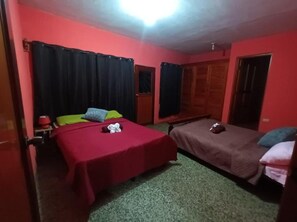 Ein Zimmer mit zwei Betten, eines mit roter Bettdecke und das andere mit brauner Bettdecke. Es gibt ein Fenster mit schwarzen Vorhängen, eine Tür und einen kleinen Tisch mit einem roten Behälter.