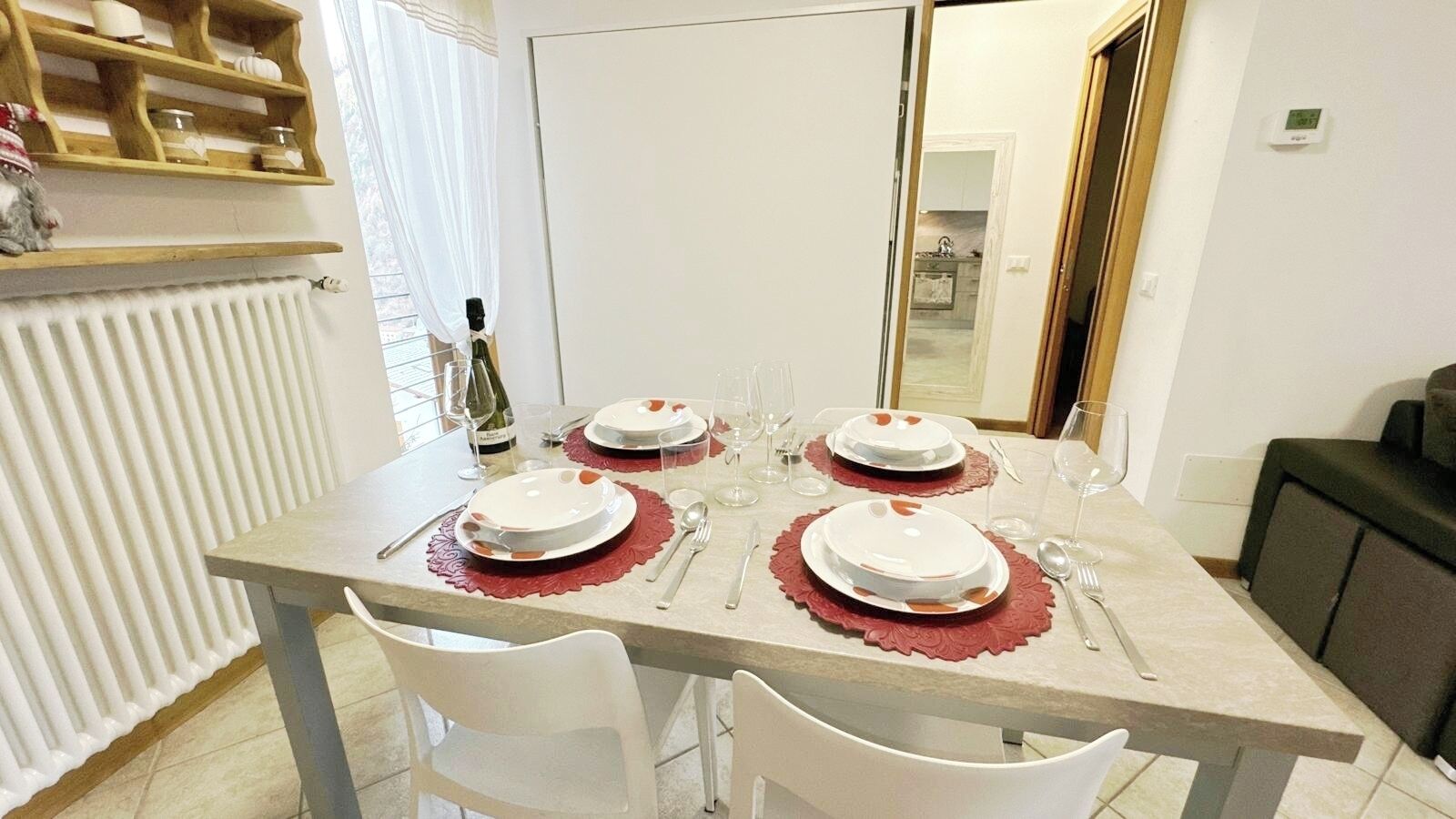 A dining table set with plates, cutlery, and a bottle of wine, with a radiator and a wooden shelf in the background.
