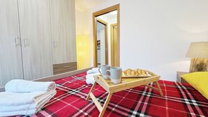 Ein Hotelzimmer mit einem Bett, einem Holzschrank, einem roten Karo-Teppich, einer Ablage mit Croissants und Tassen sowie einem gelben Kopfkissen.