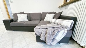 A grey sofa with two white fluffy pillows and a grey blanket on a black ottoman. - Casa Lara by Ca-Sa Check (Aprica)
