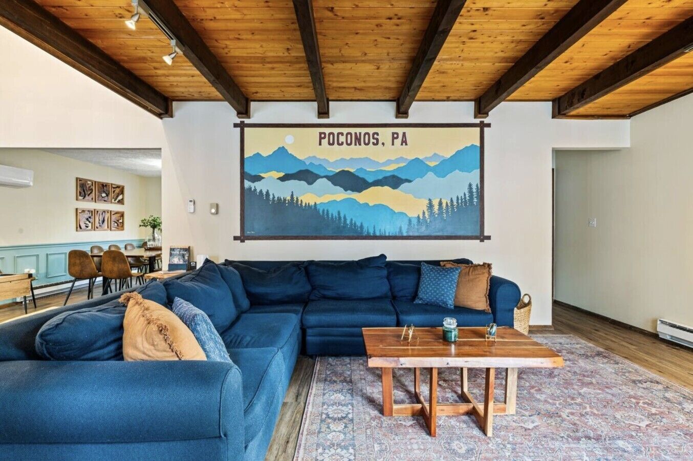 A living room with a blue sectional sofa, a wooden coffee table, and a painting of Pocono Mountains.