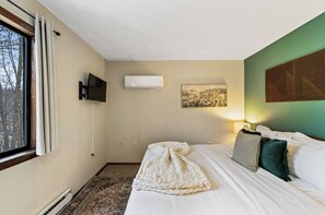 Ett hotellrum med en säng, en platt-TV, en luftkonditionering, en inramad tavla på väggen och ett fönster med gardiner.
