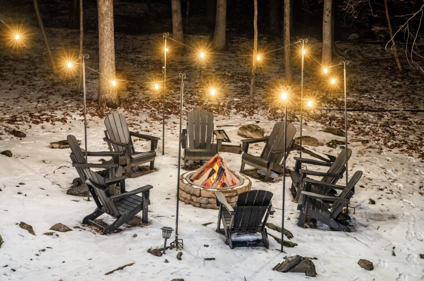 A snow-covered outdoor seating area with Adirondack chairs arranged around a central fire pit, illuminated by string lights.