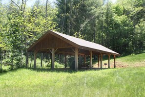 Un pavillon en bois doté d’un toit à pente, entouré d’une zone herbeuse et d’une forêt dense.