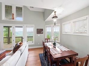 A dining area with a wooden table, chairs, and a chandelier. There are windows with blinds and a view of greenery outside. - Wintergreens R332- Mtn Condo/View, Gated Resort, Indoor/Outdoor Pools & Hot Tub (Banner Elk)