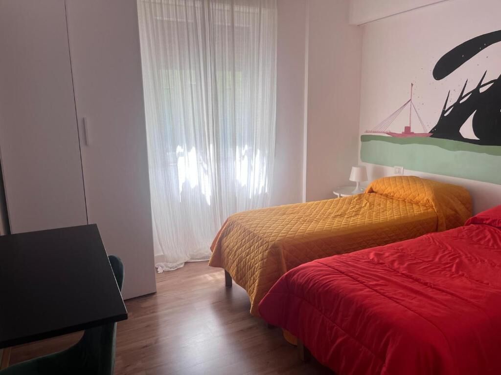 A room with two beds, a desk, and a window with sheer curtains.
