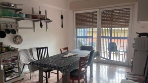 A dining area with a table, chairs, and a kitchen area with shelves and utensils.