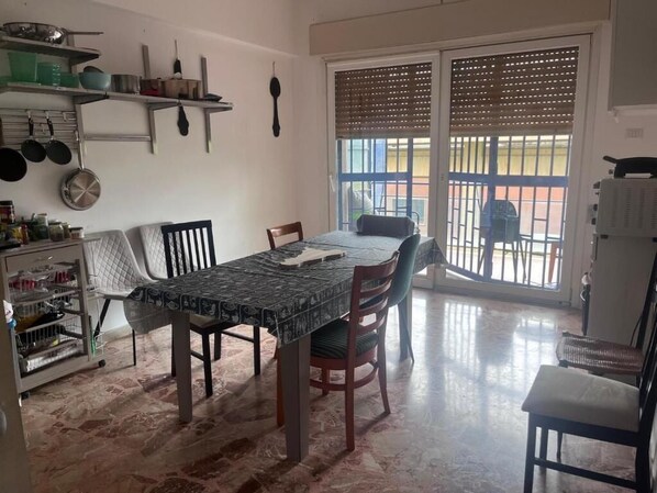 A dining area with a table, chairs, and a kitchen area with shelves and utensils.