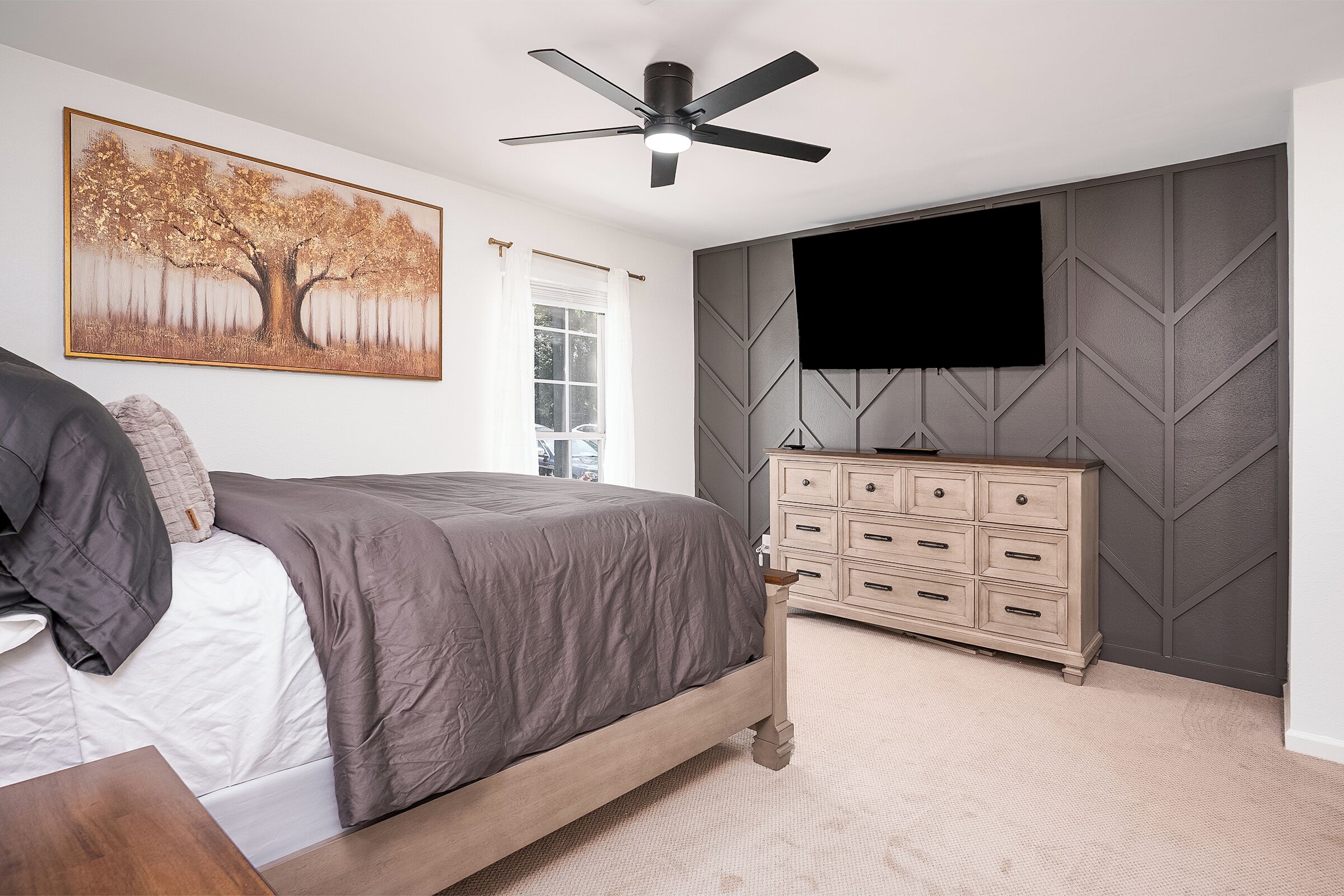 Ein Schlafzimmer mit einem Bett, einem Schminktisch, einem Deckenventilator, einem an der Wand montierten Fernseher und einem Fenster.
