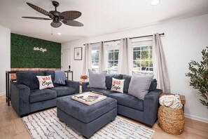 Una sala de estar moderna con un sofá modular, un ventilador de techo y vistas al exterior a través de amplias ventanas.