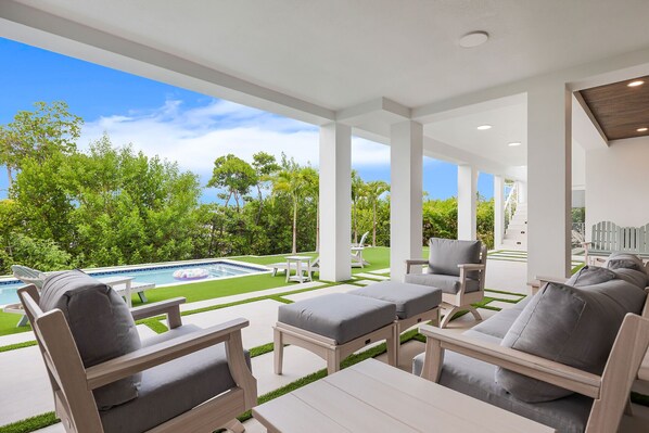 A covered outdoor seating area with a pool, white furniture, and a view of the greenery and sky. - New Luxury Key Colony Beach 5BR | Pool + 40' Dock (Key Colony Beach)