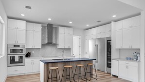 A modern kitchen with white cabinets, a central island with stools, a built-in oven, and a stainless steel refrigerator. - New Luxury Key Colony Beach 5BR | Pool + 40' Dock (Key Colony Beach)