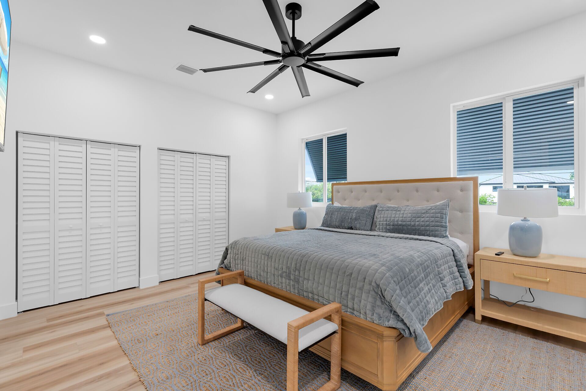 Un dormitorio con cama, mesitas de noche, un banco, ventilador de techo y ventana con persianas.
