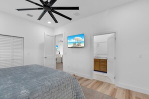 A bedroom with a bed, a ceiling fan, a TV on the wall, and a bathroom with a mirror and cabinets. - New Luxury Key Colony Beach 5BR | Pool + 40' Dock (Key Colony Beach)