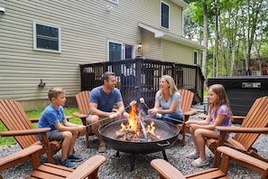 Eine Gruppe von Menschen sitzt um ein Lagerfeuer, mit einem Haus im Hintergrund.