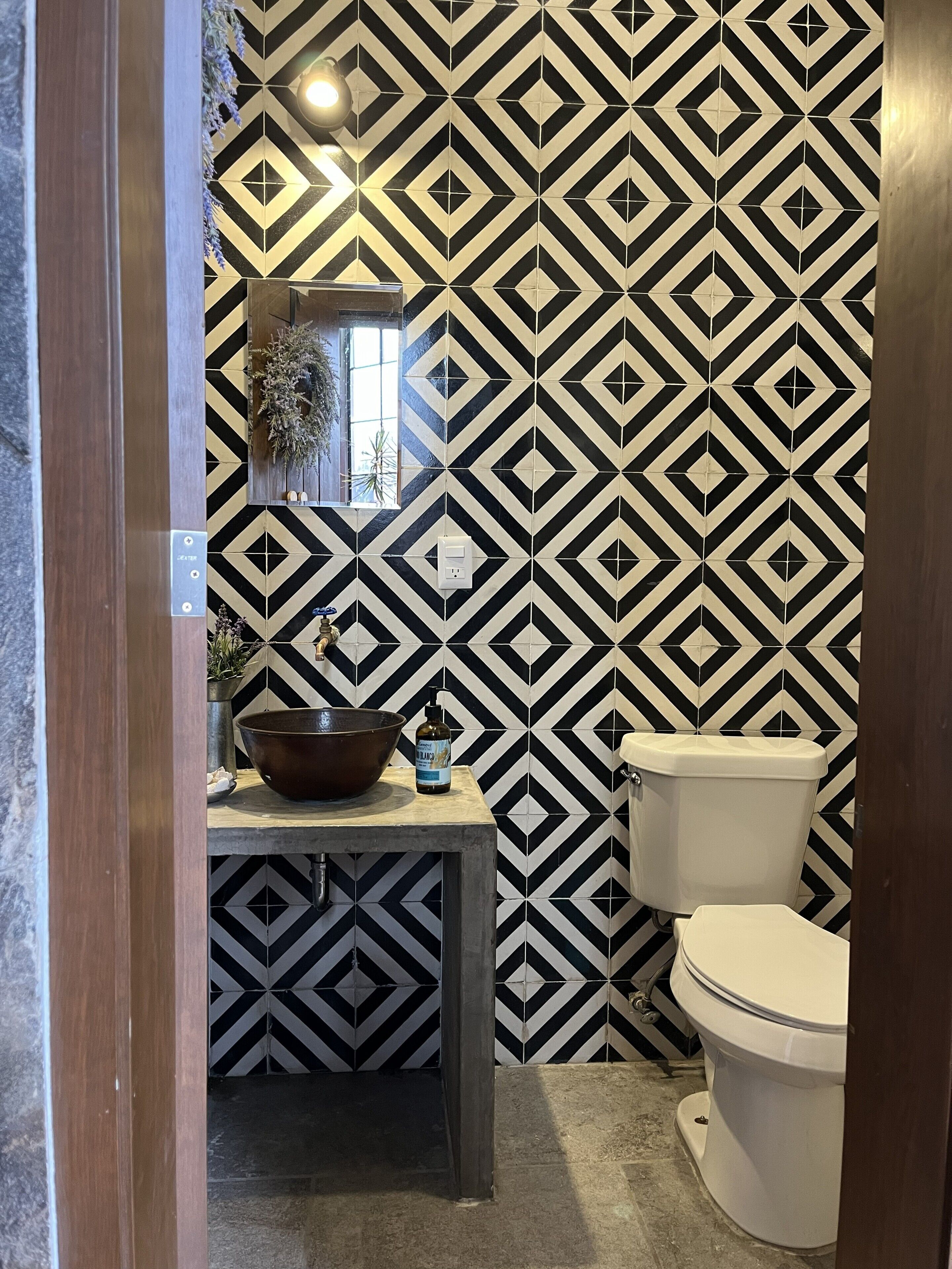A bathroom with a black and white diamond-patterned tile wall, a wooden vanity with a bowl sink, a mirror, and a toilet.