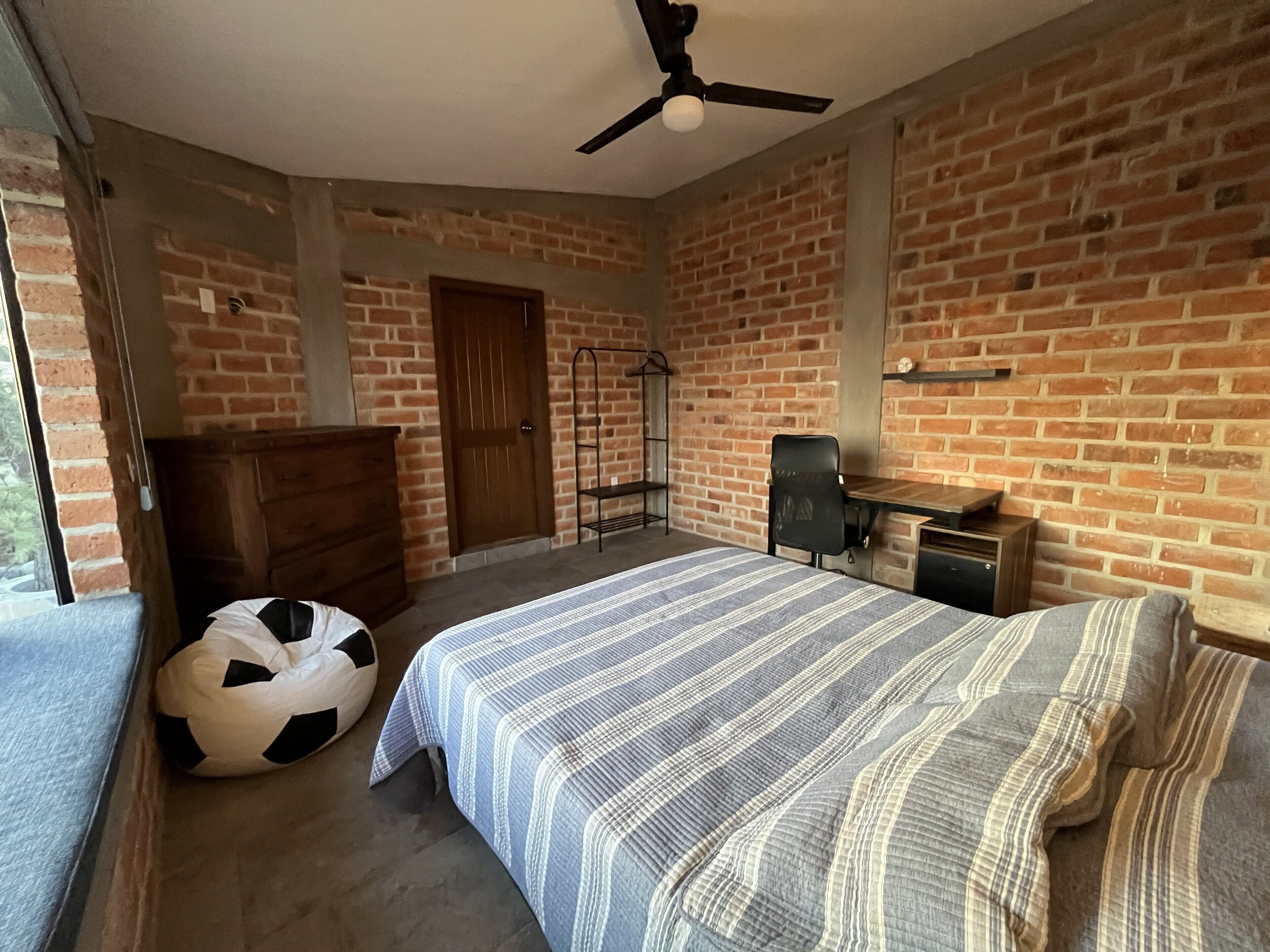 A bedroom with a brick wall, a bed with striped bedding, a wooden dresser, a black and white soccer ball, a ceiling fan, and a door.