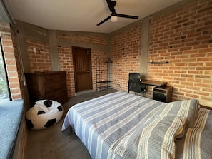 A bedroom with a brick wall, a bed with striped bedding, a wooden dresser, a black and white soccer ball, a ceiling fan, and a door.