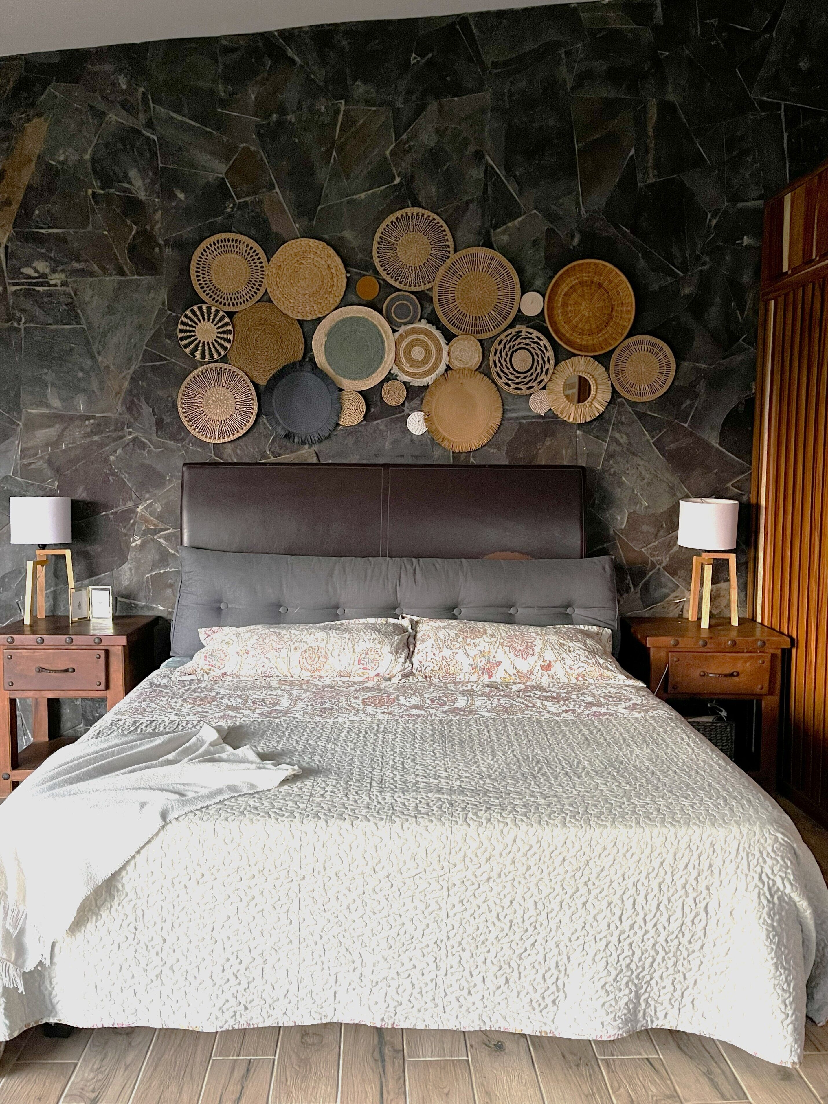 Une chambre avec un mur en pierre, un grand lit recouvert d’une courtepointe à motifs, deux tables de chevet avec des lampes et divers éléments décoratifs en tissu accrochés au mur.