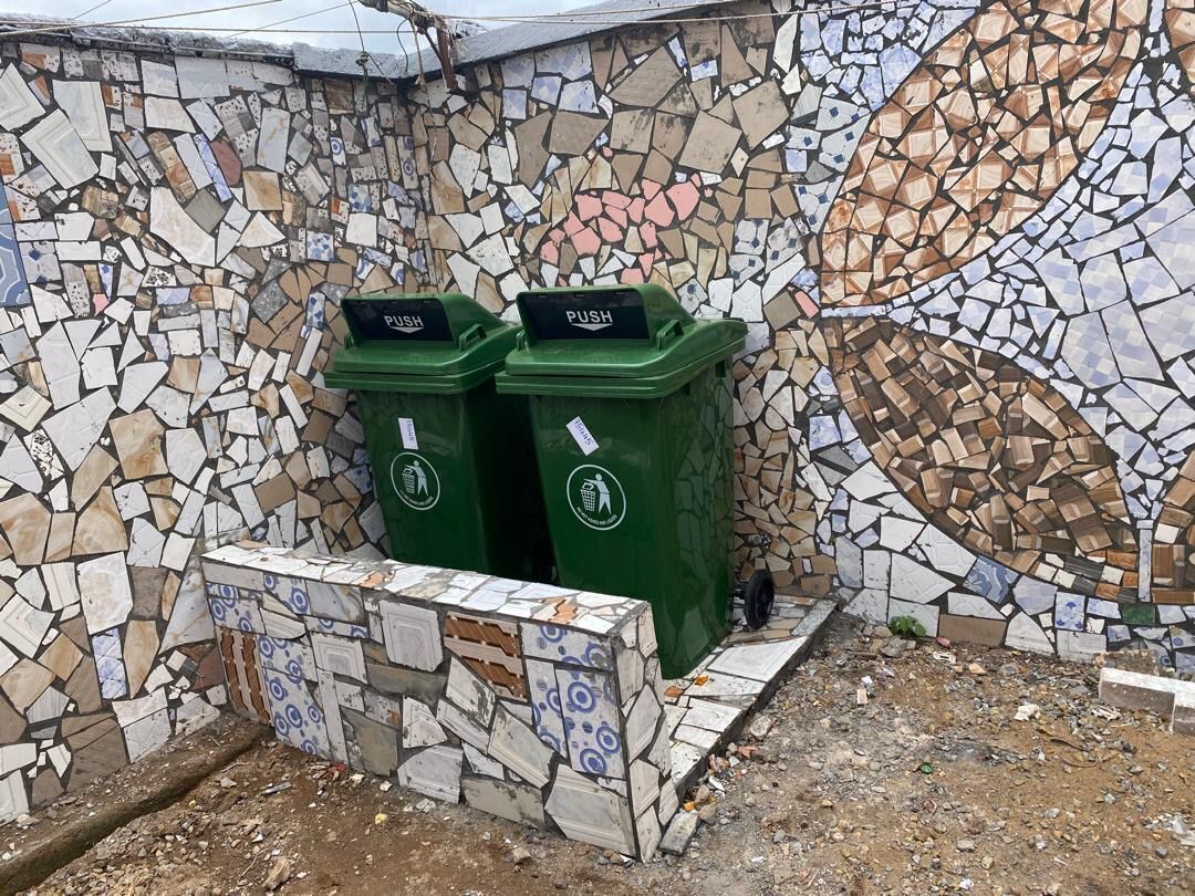 Eine Wand mit Mosaikdekor, zwei grüne Müllbehälter mit der Aufschrift 'PUSH' und eine geflieste Sitzbank.