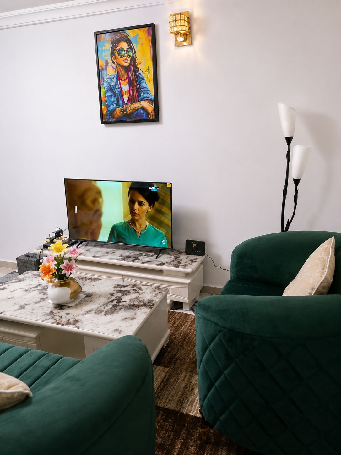 A living room with a flat-screen TV, a coffee table with a vase of flowers, and two green armchairs.