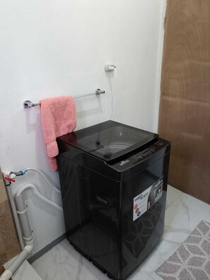 Een kleine, zwarte wasmachine met een bedieningspaneel, een roze handdoek aan een metalen rek, en een witte muur met een stopcontact.