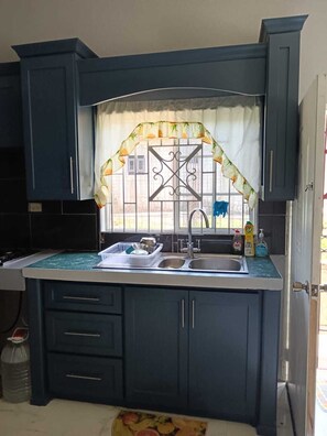 Een keuken met donkere kastjes, een raam met decoratieve gordijnen, een spoelbak met afwasmiddel en een prullenbak.