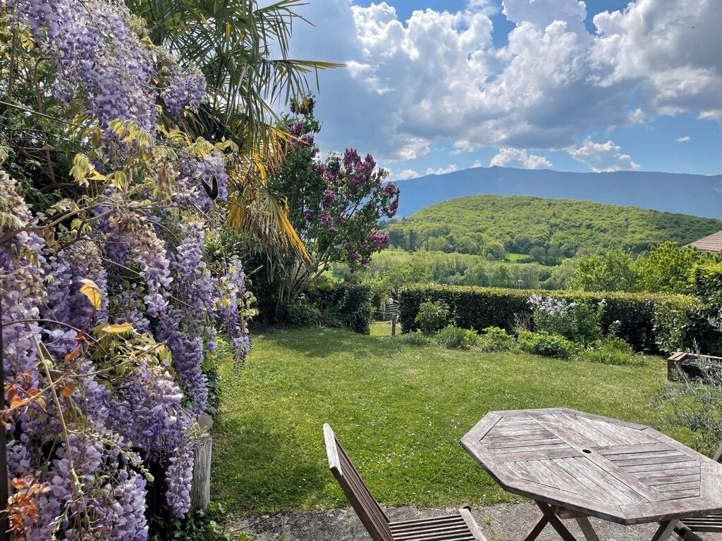 Un jardín con mesa y sillas de madera, rodeado de abundante follaje y flores púrpuras.
