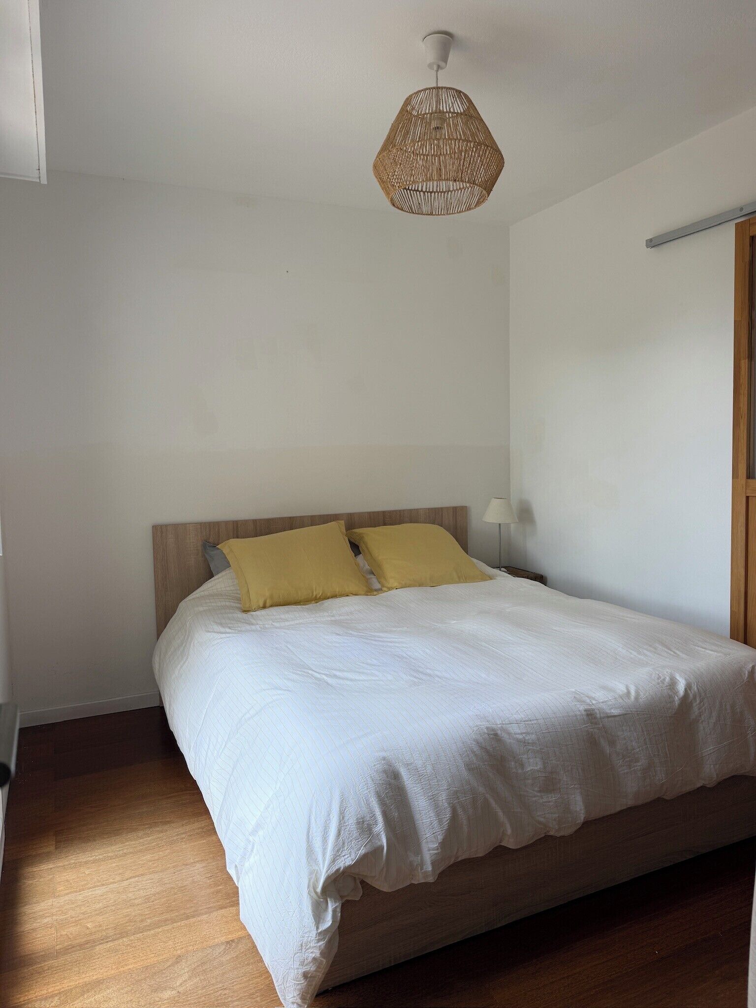 A bedroom with a wooden bed, two pillows, a bedside table with a lamp, and a woven ceiling light fixture.